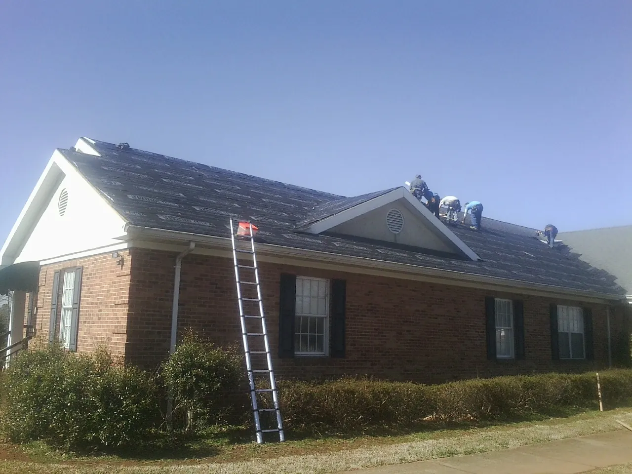 Crew tearing shingles off a brick ranch roof with a ladder set — Emergency Roof Repair work by our crew in Holly Springs