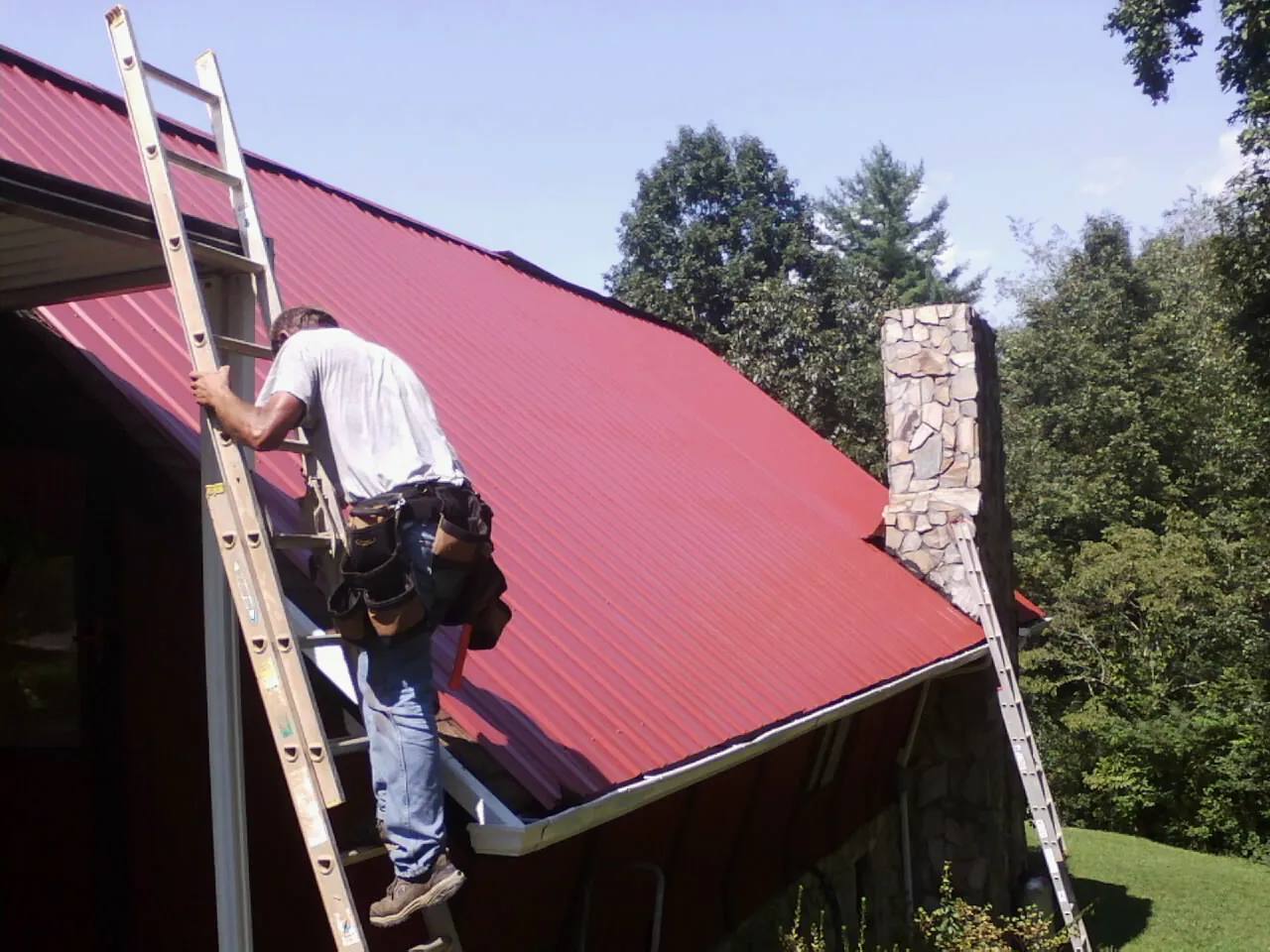 Roofer on a ladder beside a red metal roof and stone chimney — Soffit & Fascia Repair by Roofing Brothers in Holly Springs